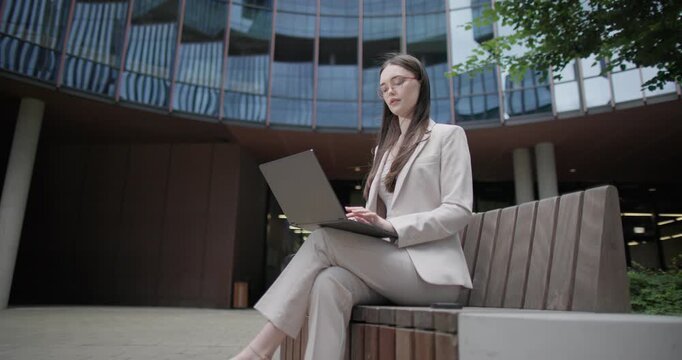 A young Caucasian businesswoman or student or intern in a business suit and glasses in an urban setting among trees and modern buildings sits on a bench and works on a laptop typing a message