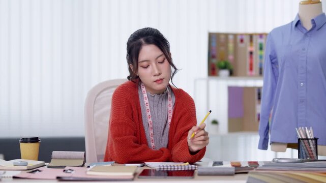 Young Asian fashion designer sketching clothing designs with measuring tape and fabric swatches in bright modern studio workspace
