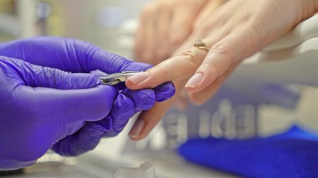 Professional manicure process in a beauty salon. Close-up of a nail technician wearing purple protective gloves carefully trimming and shaping a client's fingernail with a manicure tool.