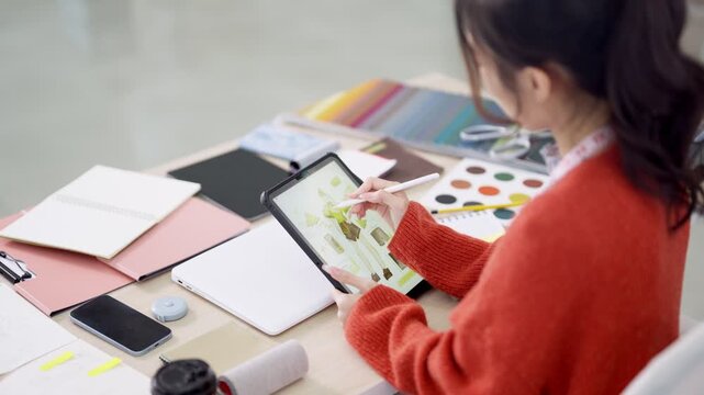 Young Asian fashion designer sketching clothing designs on digital tablet surrounded by colorful fabric swatches and material samples in bright creative studio