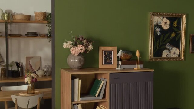 Medium long shot of modern living room interior displaying wooden cabinet with books, flower vase, and framed paintings on green wall. Clip is ideal for real estate marketing and home decor ads