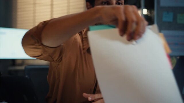 Close up overworked young Latin businessman working late on document in large stack of paper files at office night. Tax audit deadline, accounting stress, corporate burnout, urgent business concept.