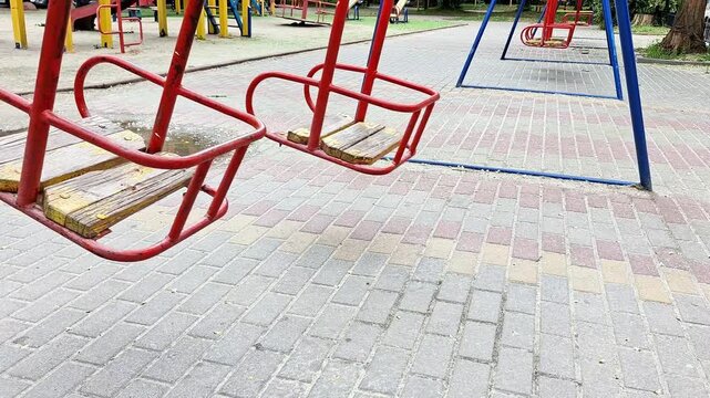 Empty red playground swing moving gently above a paved courtyard in a public park. Outdoor recreation equipment captured in daylight, symbolizing leisure, childhood, and community spaces