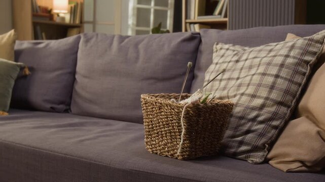 Handheld shot of knitting basket with yarn and needles resting on sofa next to plaid pillows. Clip suitable for senior lifestyle, retirement hobbies, or handcraft marketing