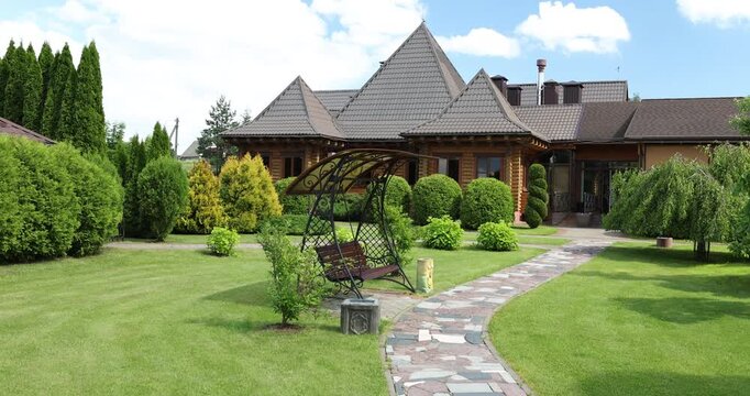 Wooden buildings with beautiful landscaped garden and green lawn. Courtyard with stone path and garden swing. View of facade of suburban residential building on sunny summer day.