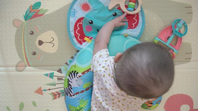 Overhead view of a baby on a soft play mat at home exploring a colorful activity pillow and toys, ideal for infant development, sensory play, childcare and parenting themes.