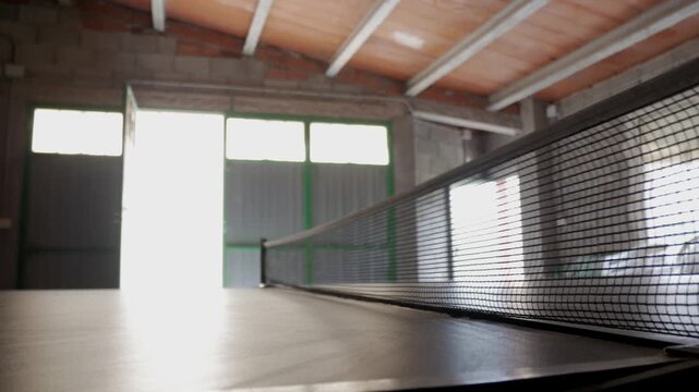 Ping pong ball bounces on table in a sunlit indoor recreational room with net visible