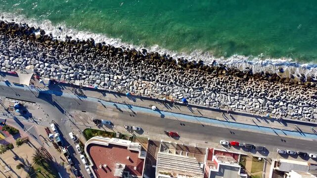 Drone aerial view looking straight down at ocean waves breaking against a coastal breakwater with tetrapods, alongside a seafront road and urban waterfront area.