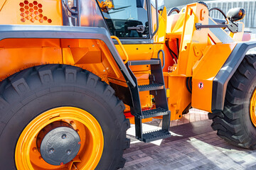 Fototapeta premium Close view of an orange construction loader showing the large front wheel, metal chassis, access steps and hydraulic components. The scene includes textured