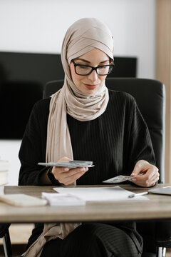 A professional woman wearing a hijab and glasses meticulously counts cash while seated at her desk, surrounded by documents and a laptop