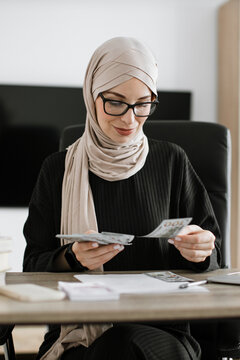 A professional woman wearing a hijab and glasses sits at a desk, meticulously counting US dollar bills, symbolizing financial management and business
