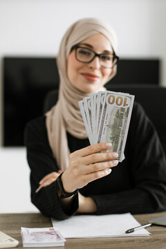 A smiling woman wearing a hijab and glasses holds a fan of US dollar bills while sitting at a desk