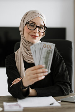A smiling woman wearing a hijab and glasses holds a fan of US dollar bills, suggesting financial success or a business transaction