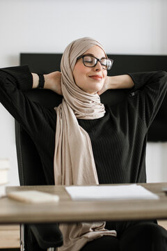 A woman wearing a hijab and glasses leans back in her chair with her hands behind her head, eyes closed in a moment of relaxation