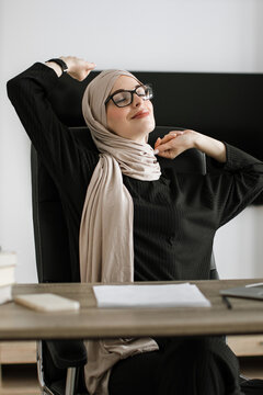 A woman wearing a hijab and glasses stretches her arms up while sitting at her desk, enjoying a moment of relaxation during her workday