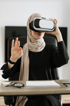 A woman wearing a hijab and VR headset gestures while seated at a desk, exploring virtual reality technology