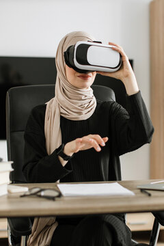 A woman wearing a hijab and a virtual reality headset is seated at a desk, engaging with digital content