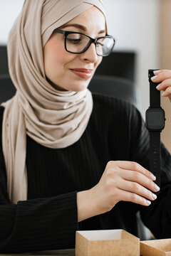 A woman wearing a hijab and glasses smiles as she holds up a new black smartwatch, ready to be worn