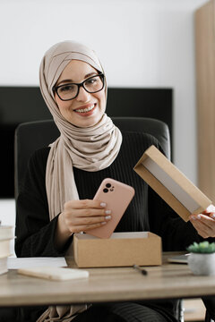 A smiling woman wearing a hijab and glasses is unboxing a new pink phone case from a cardboard box at her desk