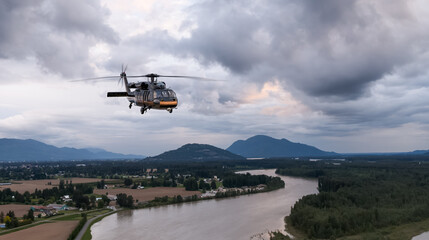 Fototapeta premium Military Helicopter Flying Over River Valley With Storm Clouds and Mountain Background in BC, Canada