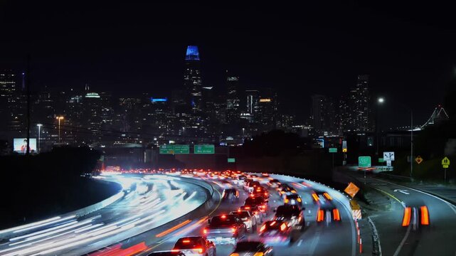 Time-lapse of heavy traffic moving along a busy highway at night, creating red and white light streaks with the illuminated San Francisco skyline, including the Salesforce Tower, in the background