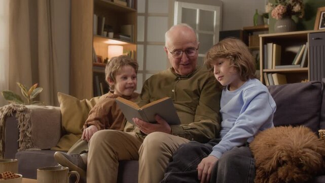 Medium long shot of senior grandfather reading book to two young grandsons sitting on sofa at home. Footage is perfect for retirement planning, family bonding, or early childhood education commercials
