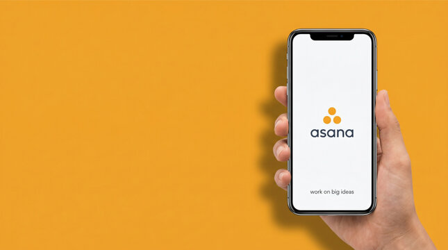 Man holding a smartphone with Asana project management application logo and tagline on the screen. Professional task tracking software mobile app interface on orange background. Mobile app mockup.
