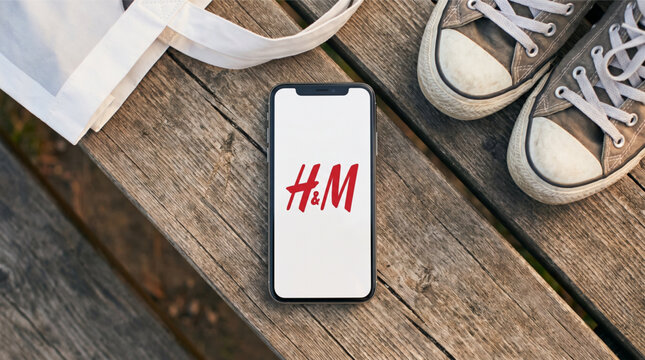 Smartphone with H&M fashion app logo on screen resting on a weathered wooden bench next to grey canvas sneakers and a white tote bag. Ideal for mobile retail and online clothing store concepts.