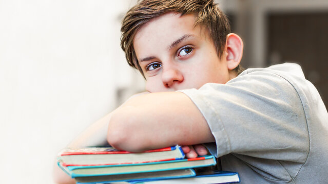 Thoughtful teenage boy lying on a stack of books tired of studying homework