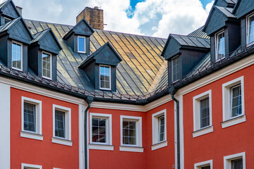 Fototapeta premium Urban architecture detail of a townhouse featuring a coral orange facade with white-framed windows and a sloped dark gray metal roof with multiple dormers, rain gutters, and a brick chimney under a cl
