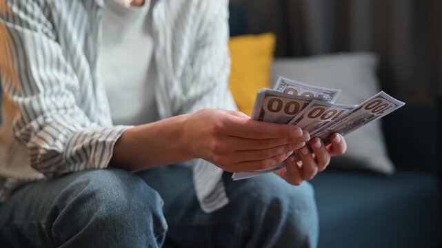 A person is seated on a couch counting a stack of cash in a comfortable indoor space. The person, who appears to be an adult female, focuses on the bills as they organize and review them