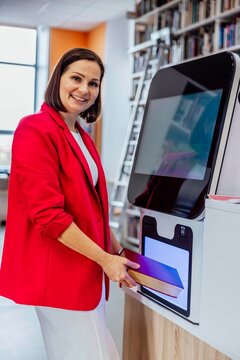 woman returning book at self service library machine looking at camera with confident slight smile in modern library interior librarian scene, education, library service