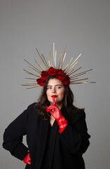 Fototapeta premium Vertical photo. Young adult white woman with DIY golden halo crown, rose on head, tulle gloves, make up, choker, collar on neck, black jacket raised index finger to red lips, looks at camera on gray