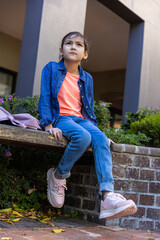 Fototapeta premium Asian girl child sitting on bench in courtyard wearing denim jacket and purple backpack beside