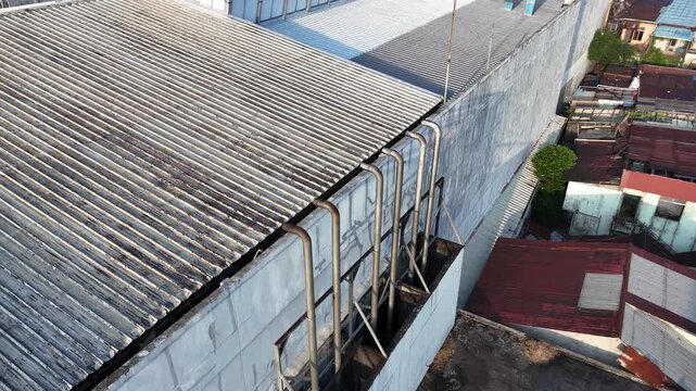 A drone shot showing a row of industrial metal exhaust pipes on the exterior wall of a factory building, with a corrugated roof in the background.