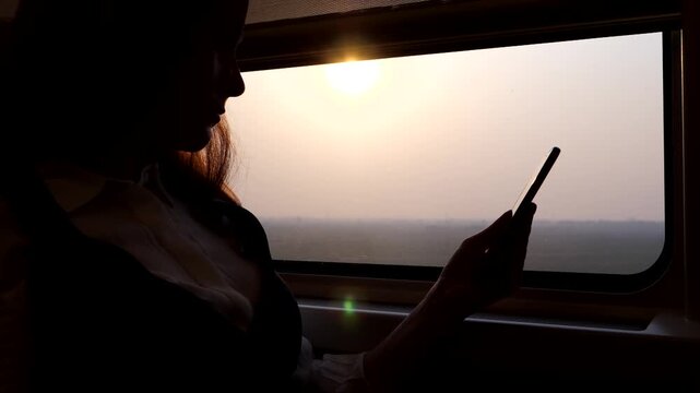 Passenger woman sit against window of high speed Chinese train, look to smartphone, slow motion silhouetted shot. Bullet train rush at Shanghai-Beijing route at evening time, bright sun shine outside