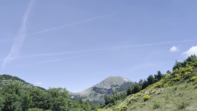 Paesaggio montano verde e luminoso, con colline erbose e un rilievo roccioso sullo sfondo sotto un cielo limpido attraversato da scie aeree
