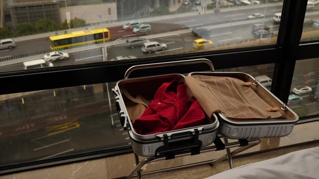 Man begin packing up, throw red shirt into open small suitcase lying against window. Slow motion shot, car traffic seen outside, down on the street. Guest prepare to leave hotel