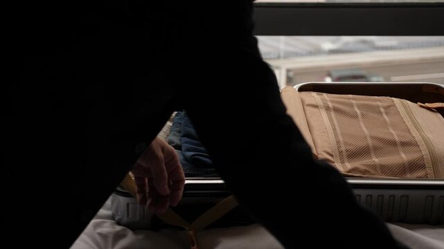 Man pack clothes into carry-on bag, place into jeans and casual shirts. Close view of open suitcase lying on bed against window. City traffic seen blurred on background