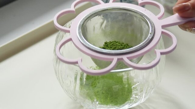 4K Close up of sifting green matcha powder through cute pink flower shaped strainer into clear glass bowl on white table, Traditional tea preparation concept.