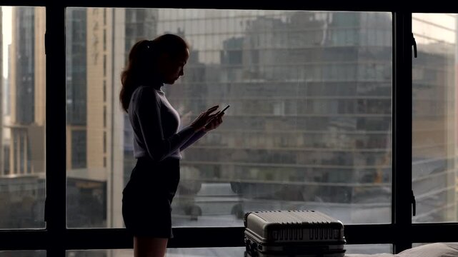 Woman stay against window at dim hotel room, using smartphone, slow motion shot. Business lady looking to mobile device and check emails. Closed suitcase lie on bed, office buildings seen outdoors