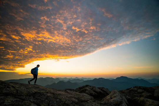Hiker walks on mountain peak against epic sunrise sky