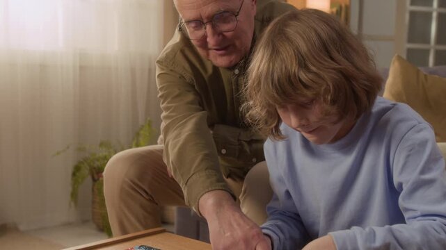 Handheld shot of Caucasian child and senior man assembling jigsaw puzzle together at home. Clip is ideal for family bonding, cognitive development, and senior care marketing campaigns