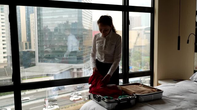 Woman take out dresses and skirts from suitcase, shake and give look, unpacking clothes at hotel room. Lady stay against window, dim interior, city buildings seen outdoors