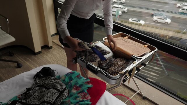 Woman continue unpacking suitcase, pick up clothes and loosely put on bed. Open trolley case lie on rack against window, city traffic seen down outdoors