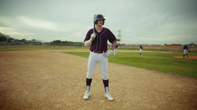 A focused baseball player in full uniform stands on the dirt infield, bat resting on his shoulder, preparing for the next play. Teammates are in the background on the green outfield.