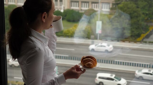 Woman stand still against window, make sip of coffee from cup, hold small plate with croissant. Lady look out to city, car traffic seen down outside on the road