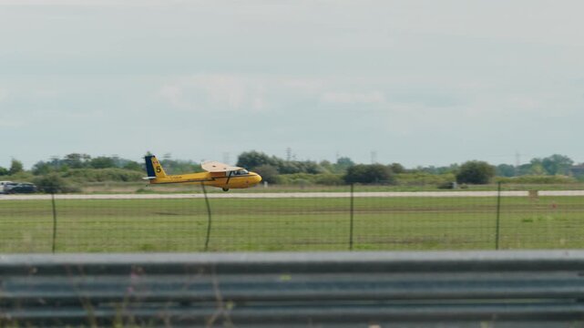 Tracking shot of a yellow glider plane landing in a grassy field beside a local country airstrip, showing a calm rural aviation scene.
