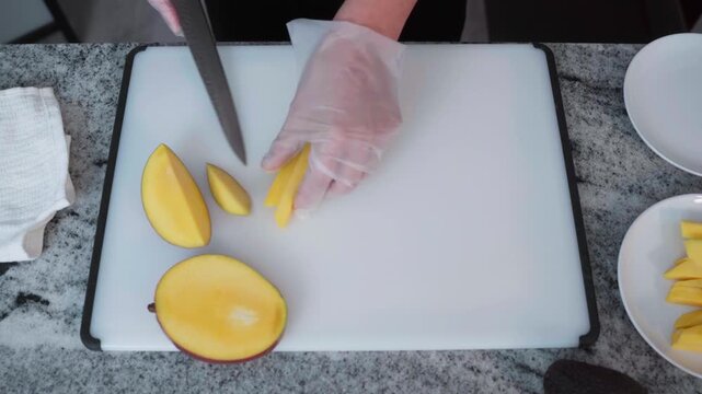 An overhead shot of the kitchen shows gloved hands slicing a ripe mango into thin slices on a white cutting board.