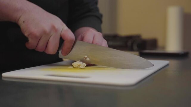 A close-up kitchen shot shows hands mincing or chopping garlic on a white cutting board with a large chef&rsquo;s knife.